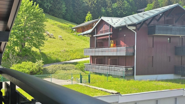 A multi-story chalet with balconies, set in a mountainous area with a grassy slope and a road. - Studio on hiking trails and paths.  Ideal for a couple. (Les Gets)