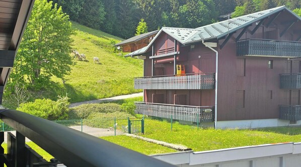 A multi-story chalet with balconies, set in a mountainous area with a grassy slope and a road. - Studio on hiking trails and paths.  Ideal for a couple. (Les Gets)