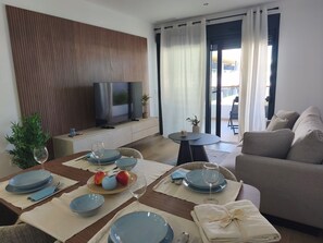 A modern living room with a flat-screen TV, a sofa, a dining table set for four, and a balcony with a view. - Lagoons Premium Apartment (Alicante)