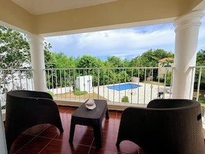 A balcony with two wicker chairs and a small table, overlooking a swimming pool and lush greenery.