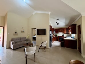 A spacious living area with a sofa, chairs, a TV, and a kitchen with wooden cabinets and a refrigerator.