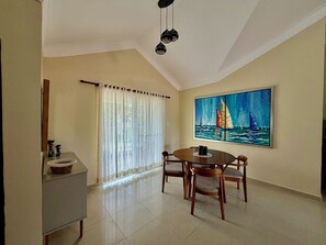 Un coin repas avec une table ronde et des chaises, un tableau représentant des voiliers accroché au mur et une fenêtre ornée de rideaux légers.