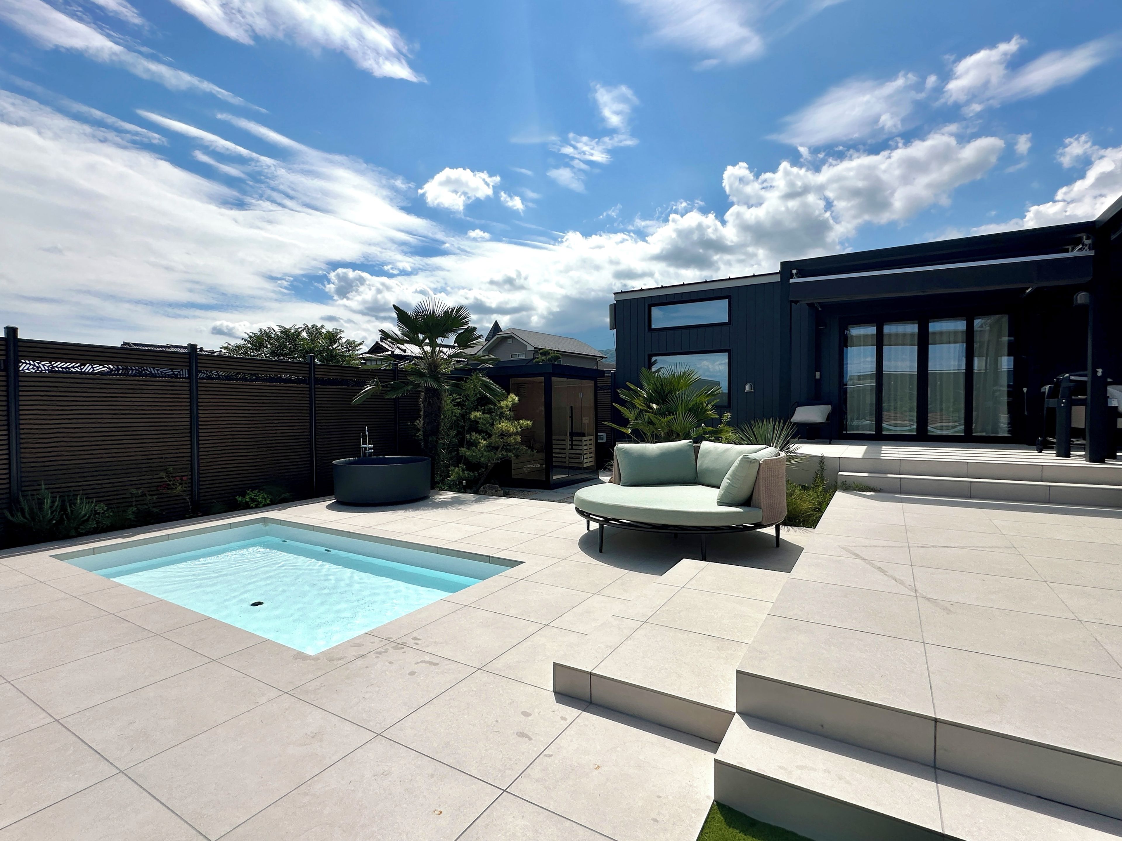A modern outdoor pool area with a built-in hot tub, a light-colored sofa, and a contemporary building with large windows.