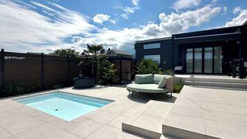 A modern outdoor pool area with a built-in hot tub, a light-colored sofa, and a contemporary building with large windows.