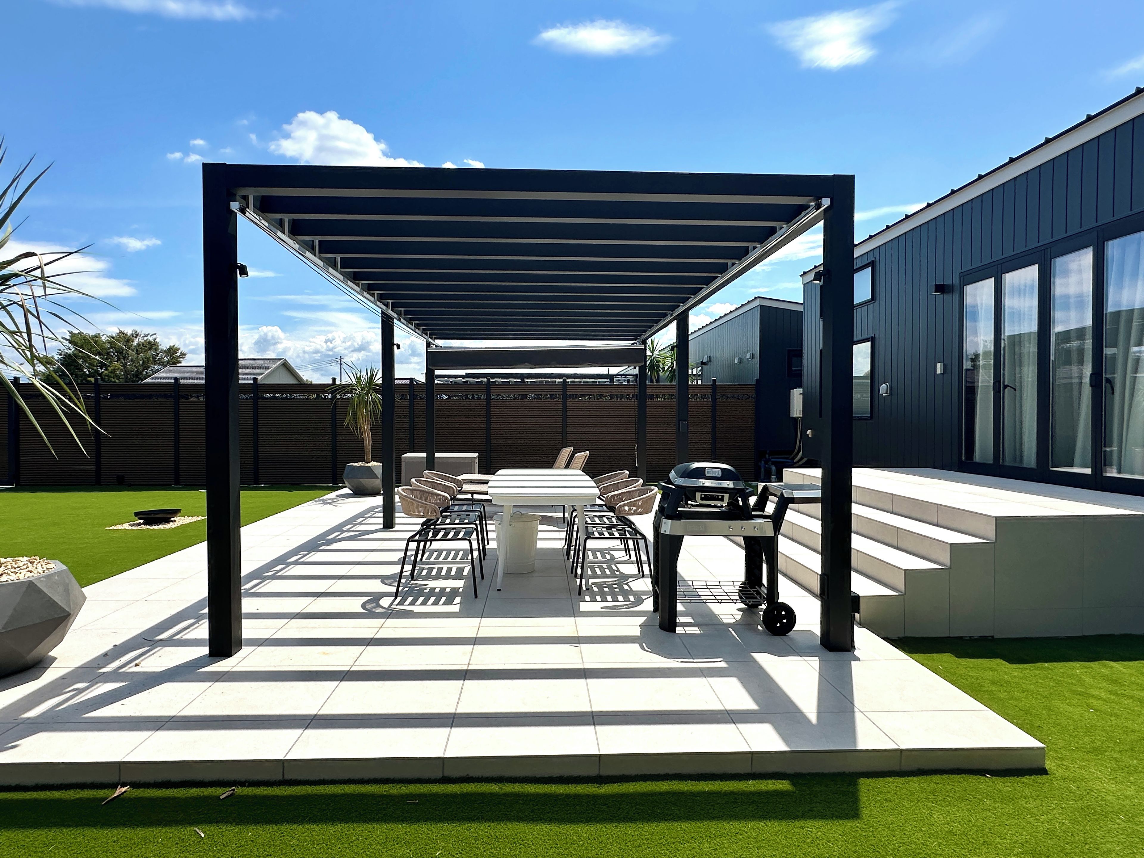 A covered patio area with a table and chairs, a grill, and a modern building in the background.