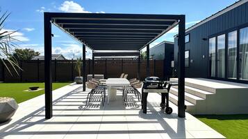 A covered patio area with a table and chairs, a grill, and a modern building in the background.