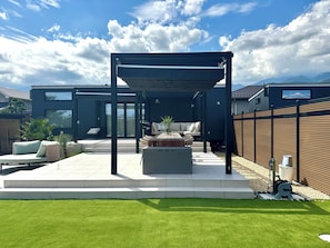 Eine moderne Terrassenanlage mit überdachter Sitzgruppe, Feuerstelle und gepflegtem Rasen.
