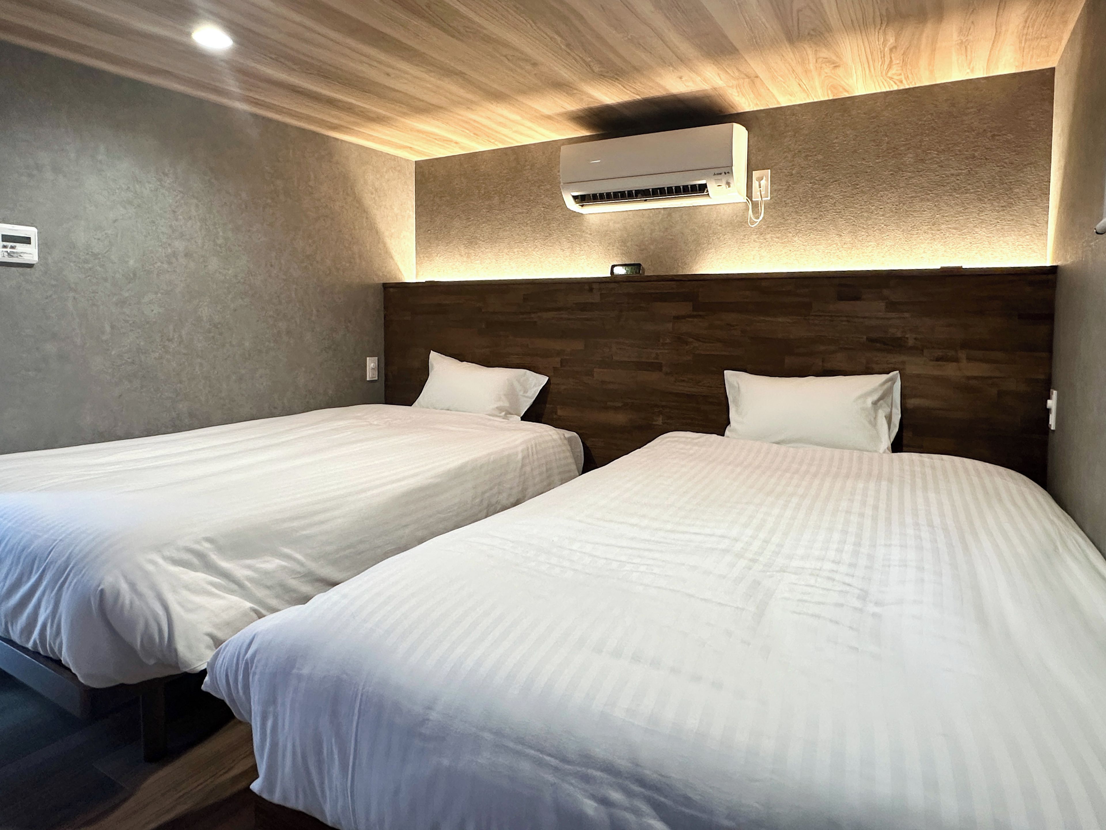 A hotel room with two single beds, a wooden headboard, an air conditioning unit, and a wall-mounted thermostat.