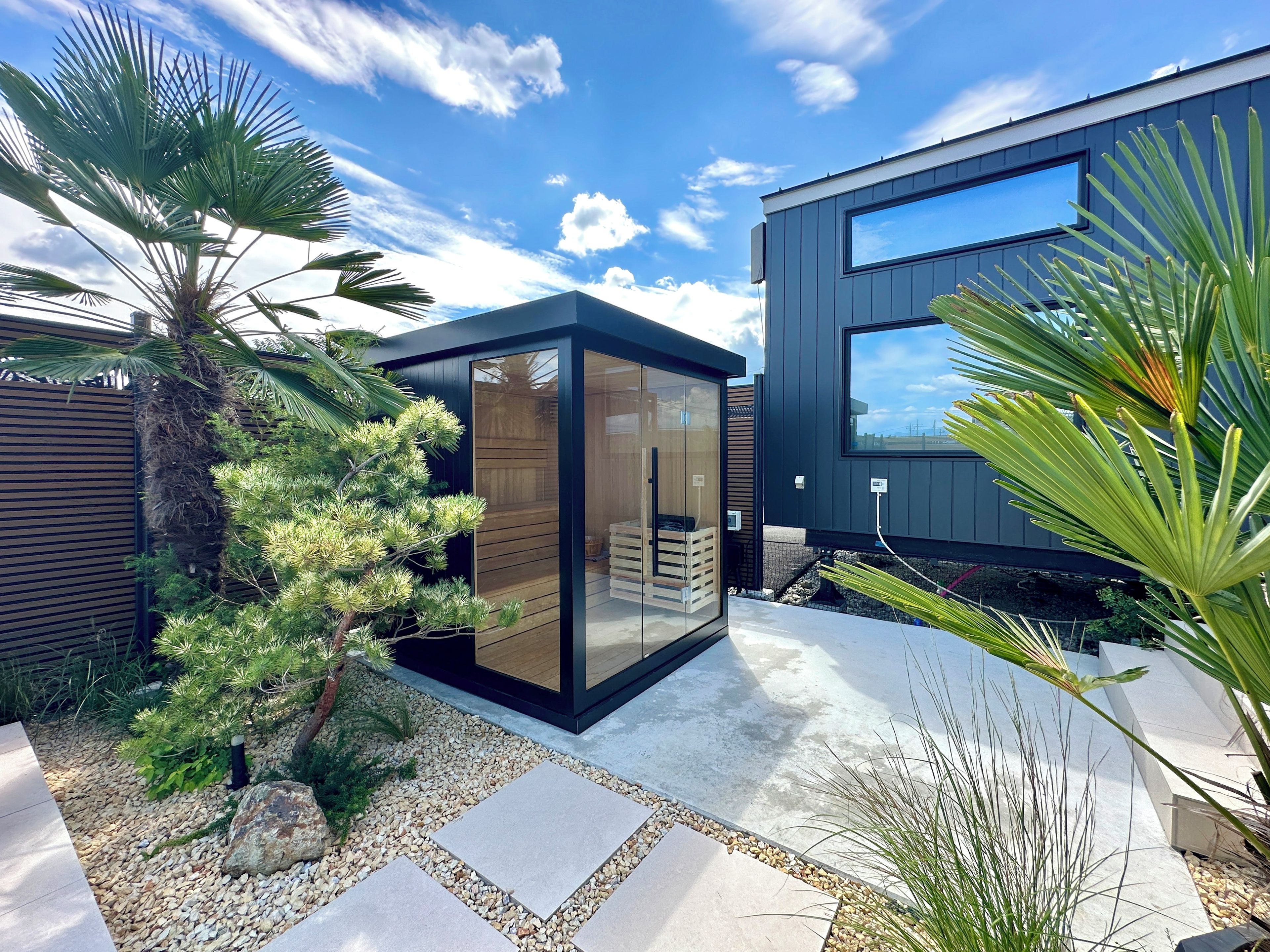 Un bâtiment moderne doté d’une façade en verre, entouré d’un jardin planté de palmiers et traversé par un sentier de gravier.