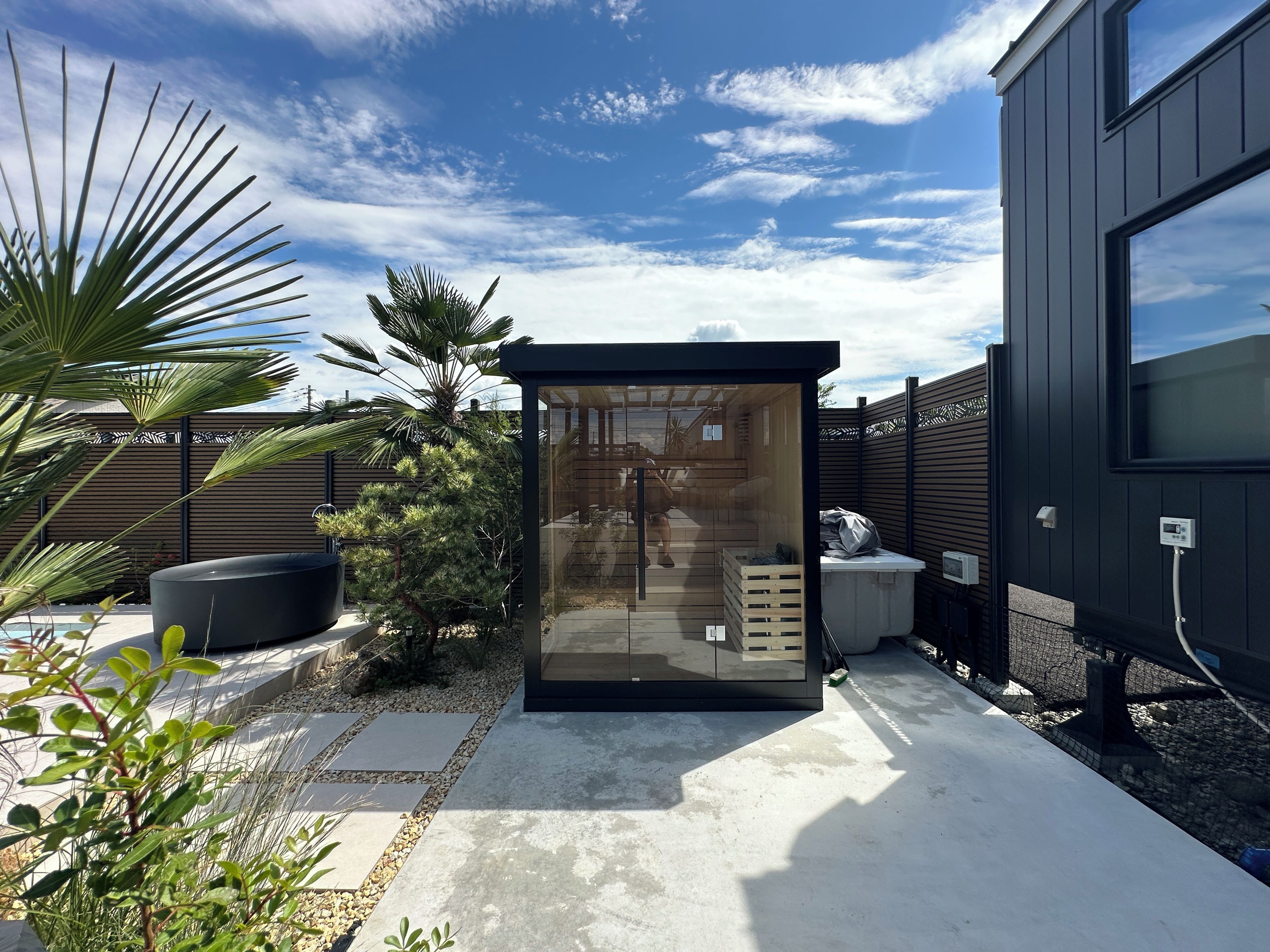 A modern outdoor structure with a glass door, surrounded by a garden with various plants and a wooden fence.