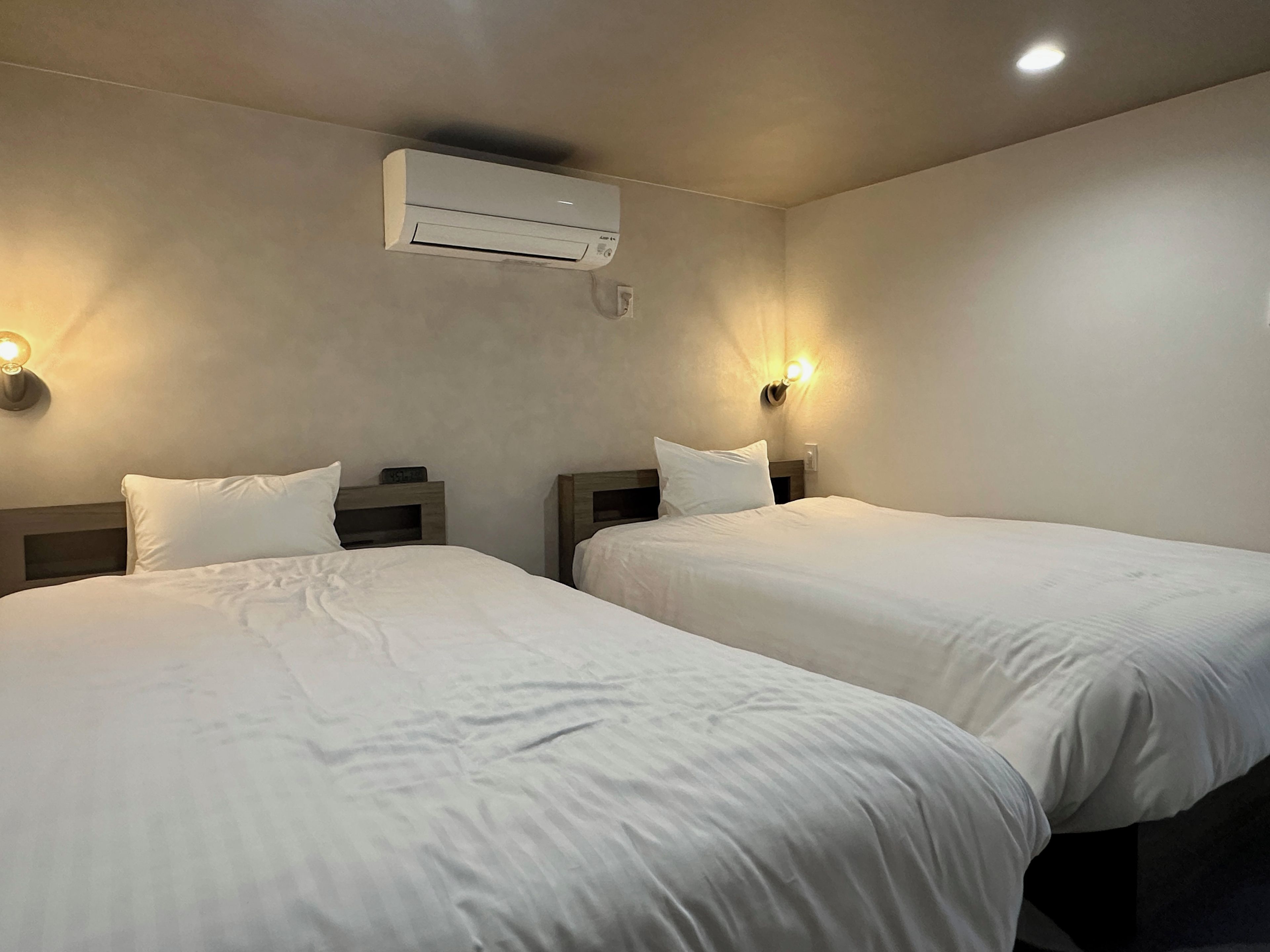 Ein Hotelzimmer mit zwei Betten, einer Wand-Klimaanlage und Nachttischlampen.