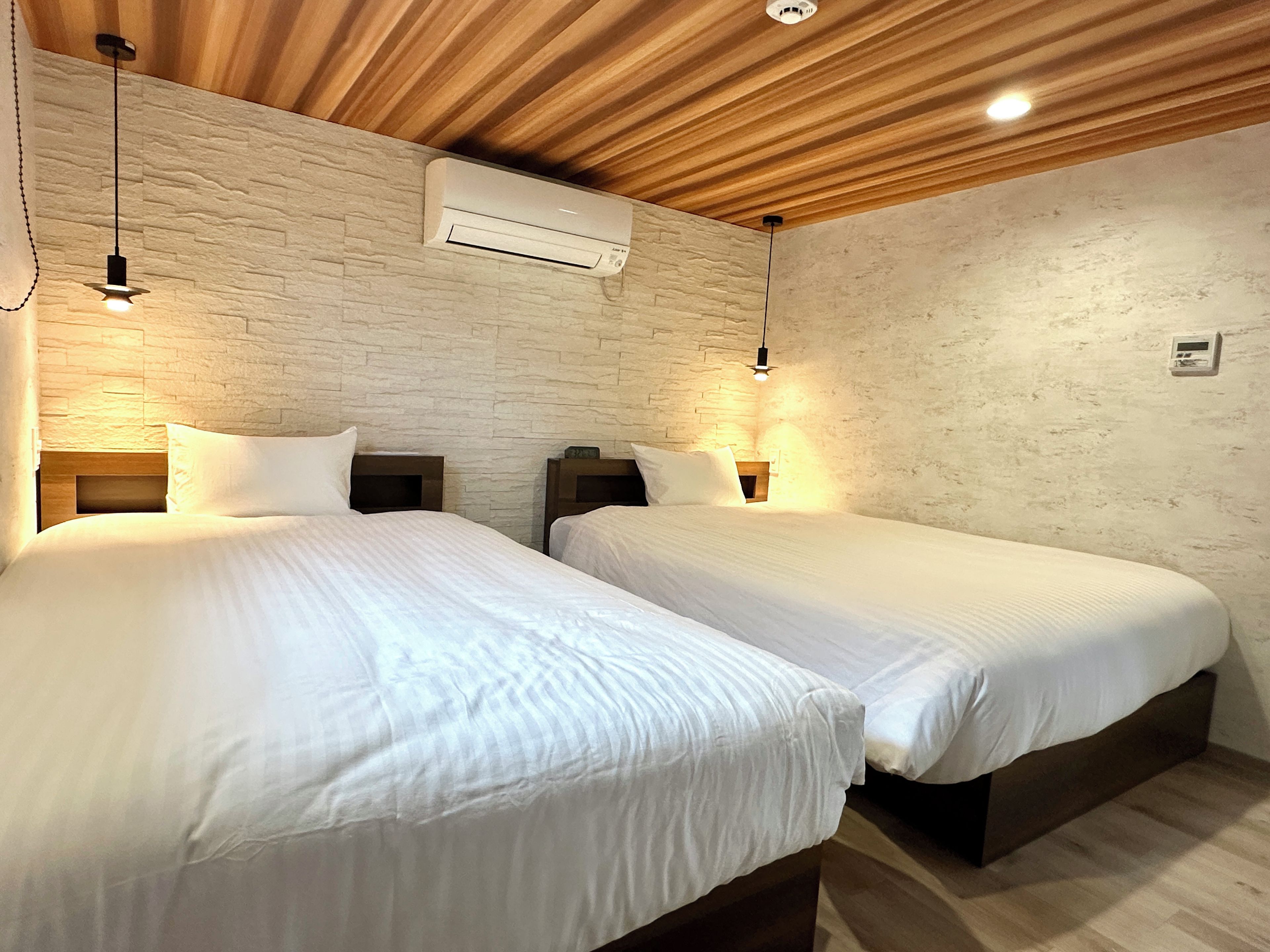 Habitación de hotel con dos camas, techo de madera, pared de piedra y aire acondicionado instalado en el techo.