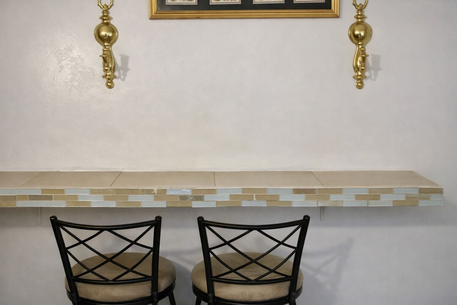 Une cuisine avec un mur recouvert de carreaux, deux chaises noires et un tableau encadré au-dessus du comptoir.