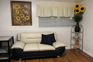 Een woonkamer met een bank, een tafel, een raam met jaloezieën, een lijst met een afbeelding en een vaas met bloemen.
