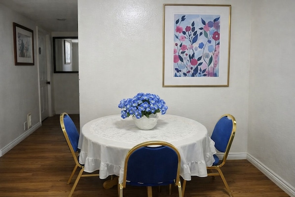 A dining room with a round table, blue chairs, a vase of flowers, and a framed floral painting on the wall.