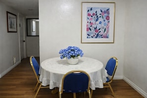Een eetkamer met een ronde tafel, blauwe stoelen, een bloevas en een lijst met een bloemistisch schilderij aan de muur.