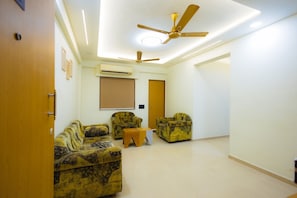 Um quarto com ventilador de teto, um sofá, duas poltronas, uma mesinha e um aparelho de ar condicionado.