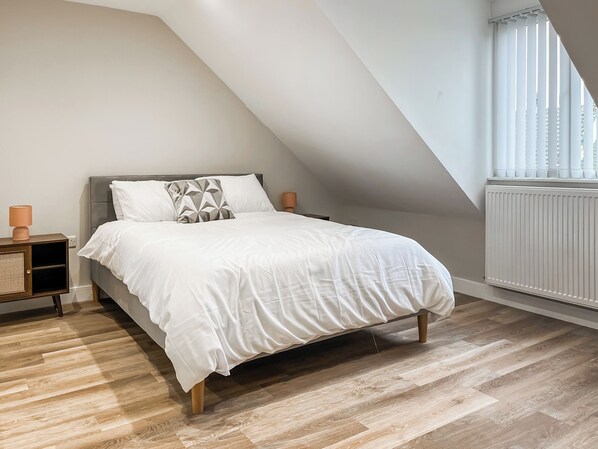 A bedroom with a sloped ceiling, a bed with white bedding, a bedside table with a lamp, and a window with blinds.