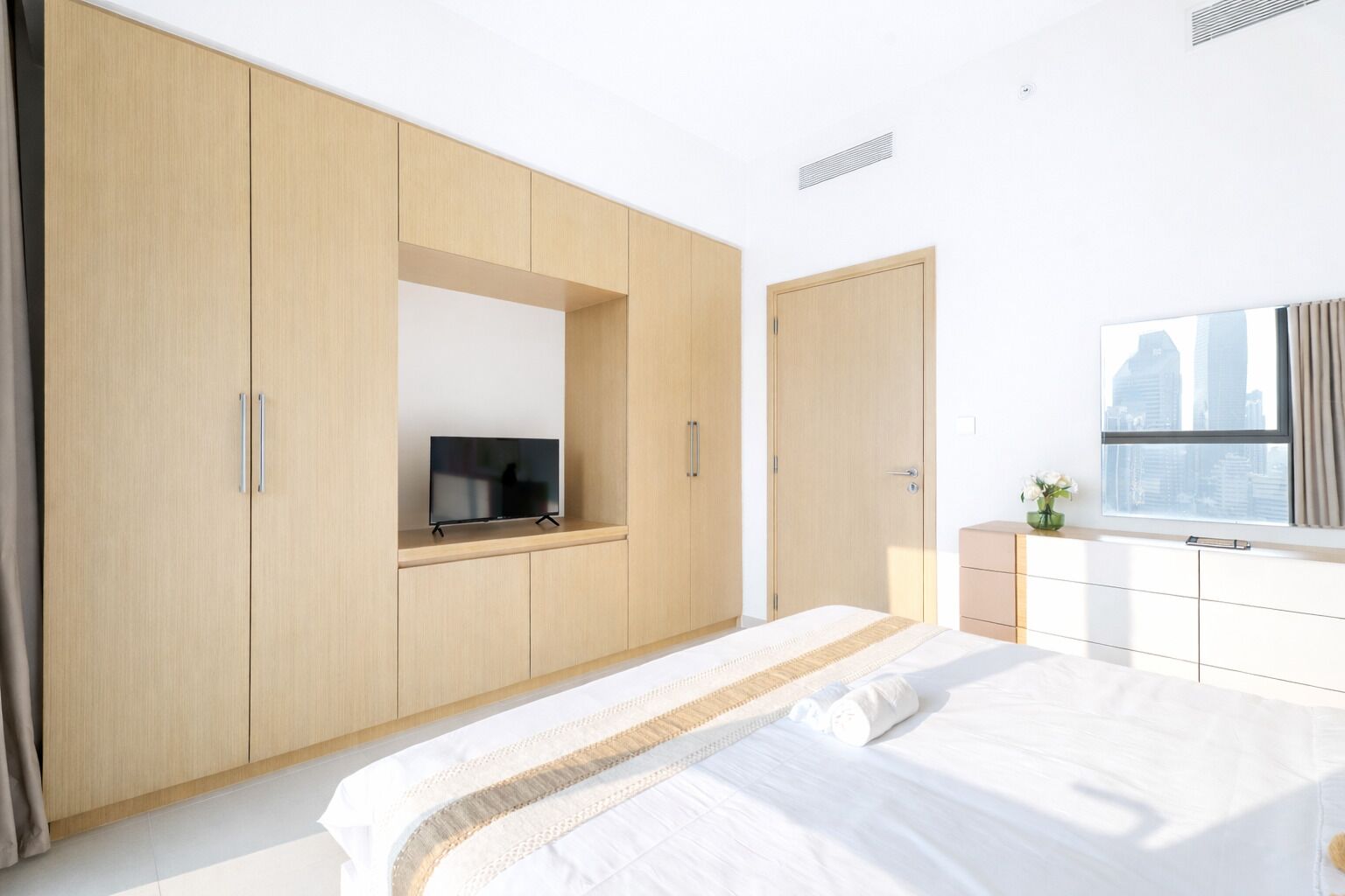 A modern bedroom with a large bed, a TV mounted on a wooden cabinet, and a window with a city view.