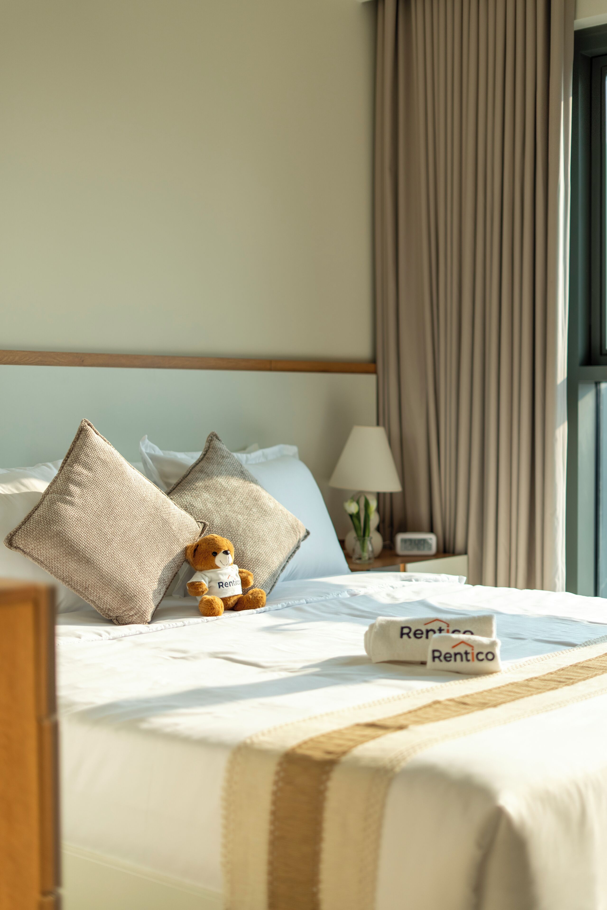 A neatly made bed with a pillow, a small teddy bear, a lamp, and a 'Rentico' branded towel.