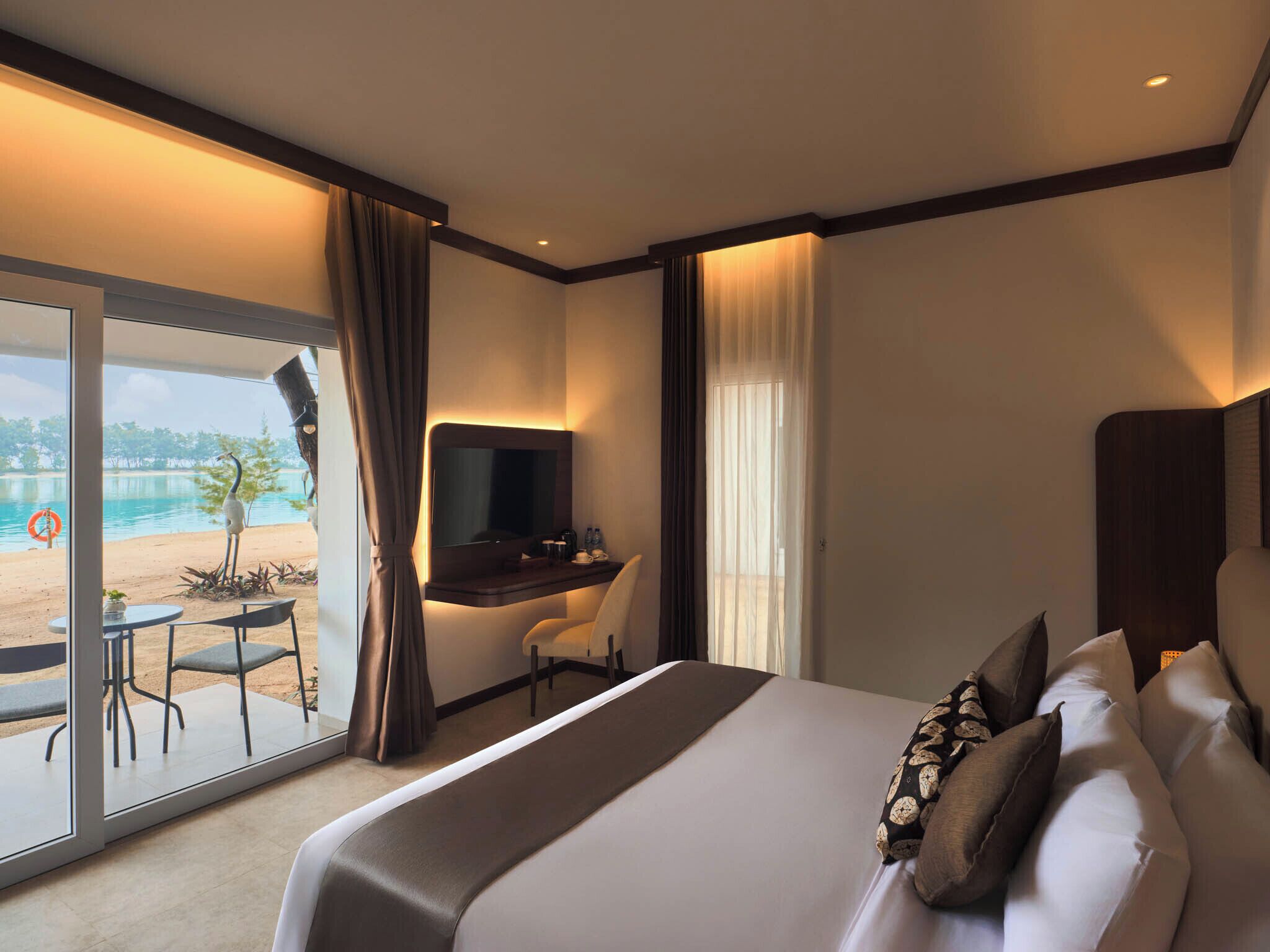 A hotel room with a bed, a desk, a chair, a TV, and a view of the beach.