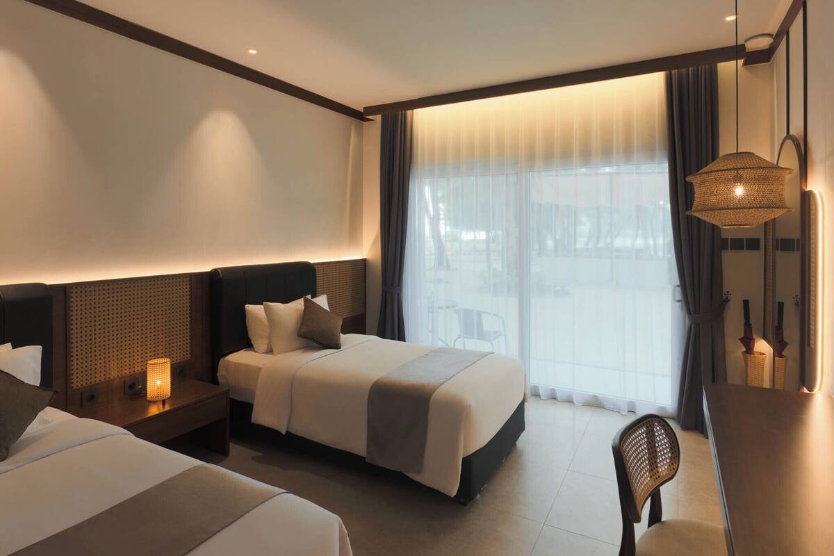 Ein Hotelzimmer mit zwei Betten, einem Schreibtisch, einem Stuhl, einem großen Fenster mit Vorhängen und einer Hängelampe.