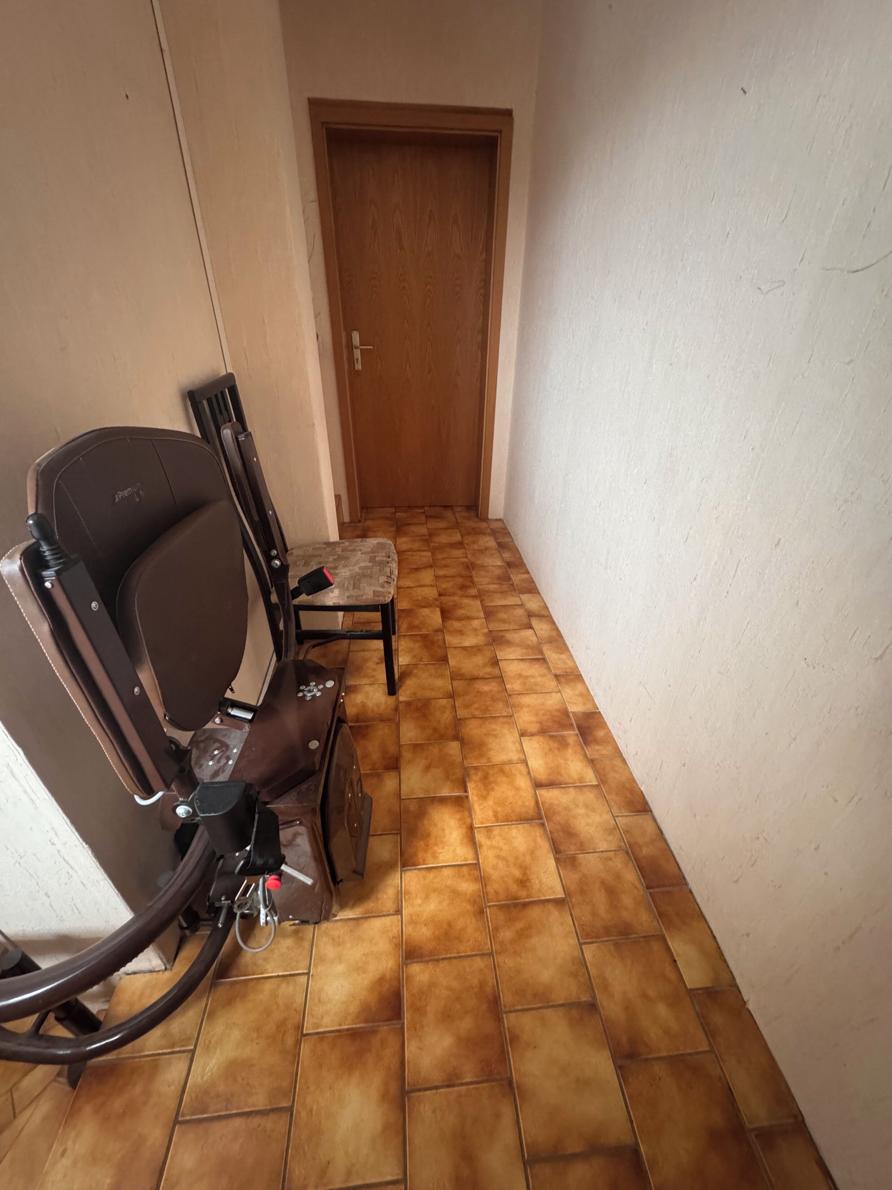 Un pasillo estrecho con una puerta de madera al final, un sillón de cuero marrón y un taburete pequeño.