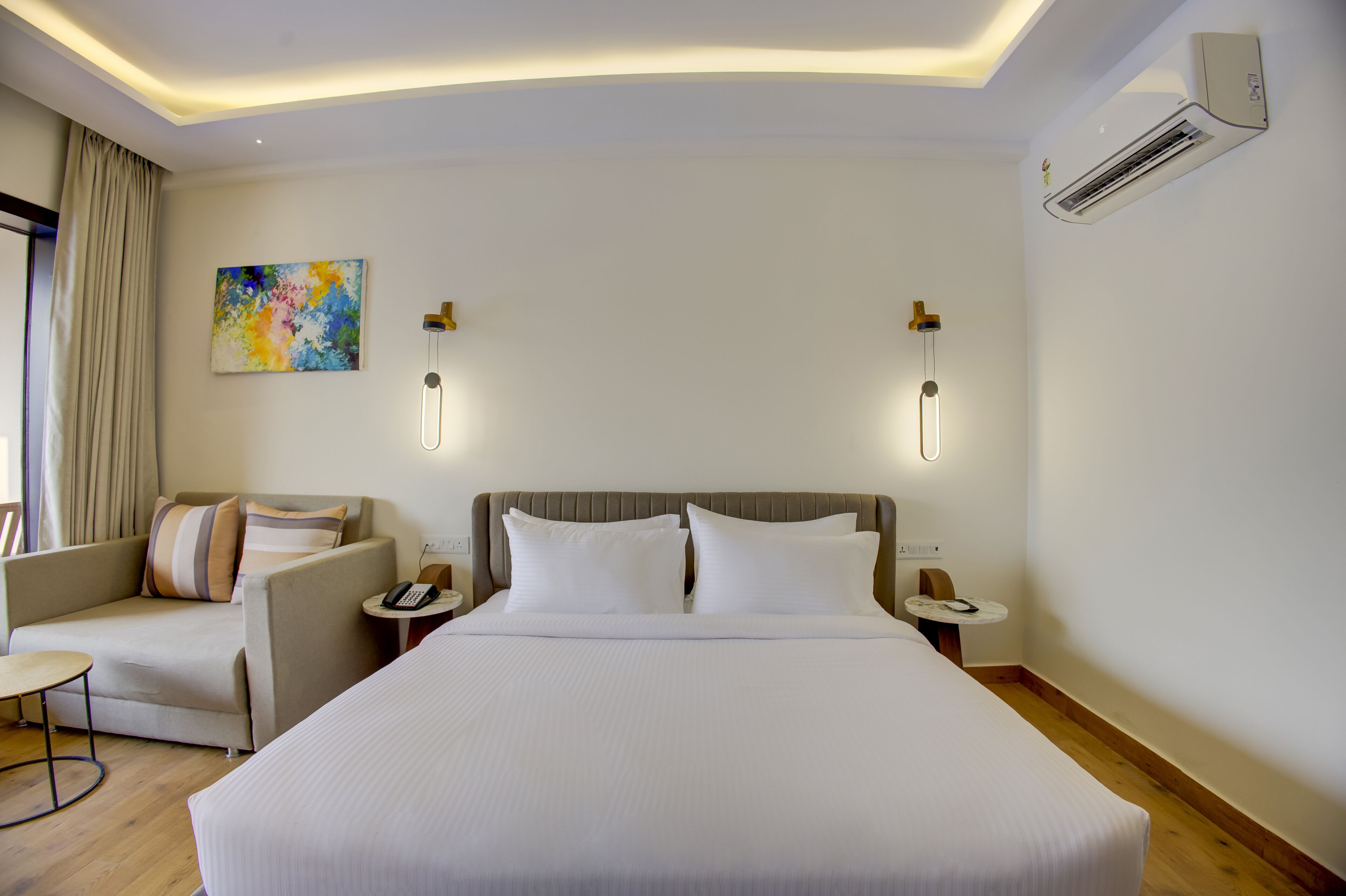 A hotel room with a large bed, a sofa, a small table, and an air conditioning unit.