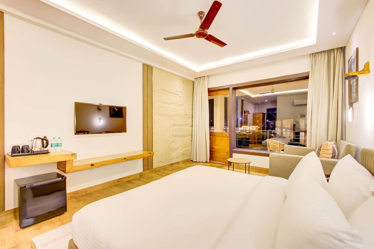 A hotel room with a bed, a TV, a desk with a coffee maker, a small table, and a view of the hotel lobby.