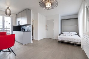 A modern hotel room with a bed, a red chair, a kitchenette, and a large window. - Eiffel Tower View Studio w/ Balcony & Elevator (Paris)