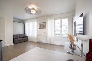 A modern living room with a grey sofa, a white cabinet, a framed abstract painting, and a large window with white curtains. - Eiffel Tower View Studio w/ Balcony & Elevator (Paris)