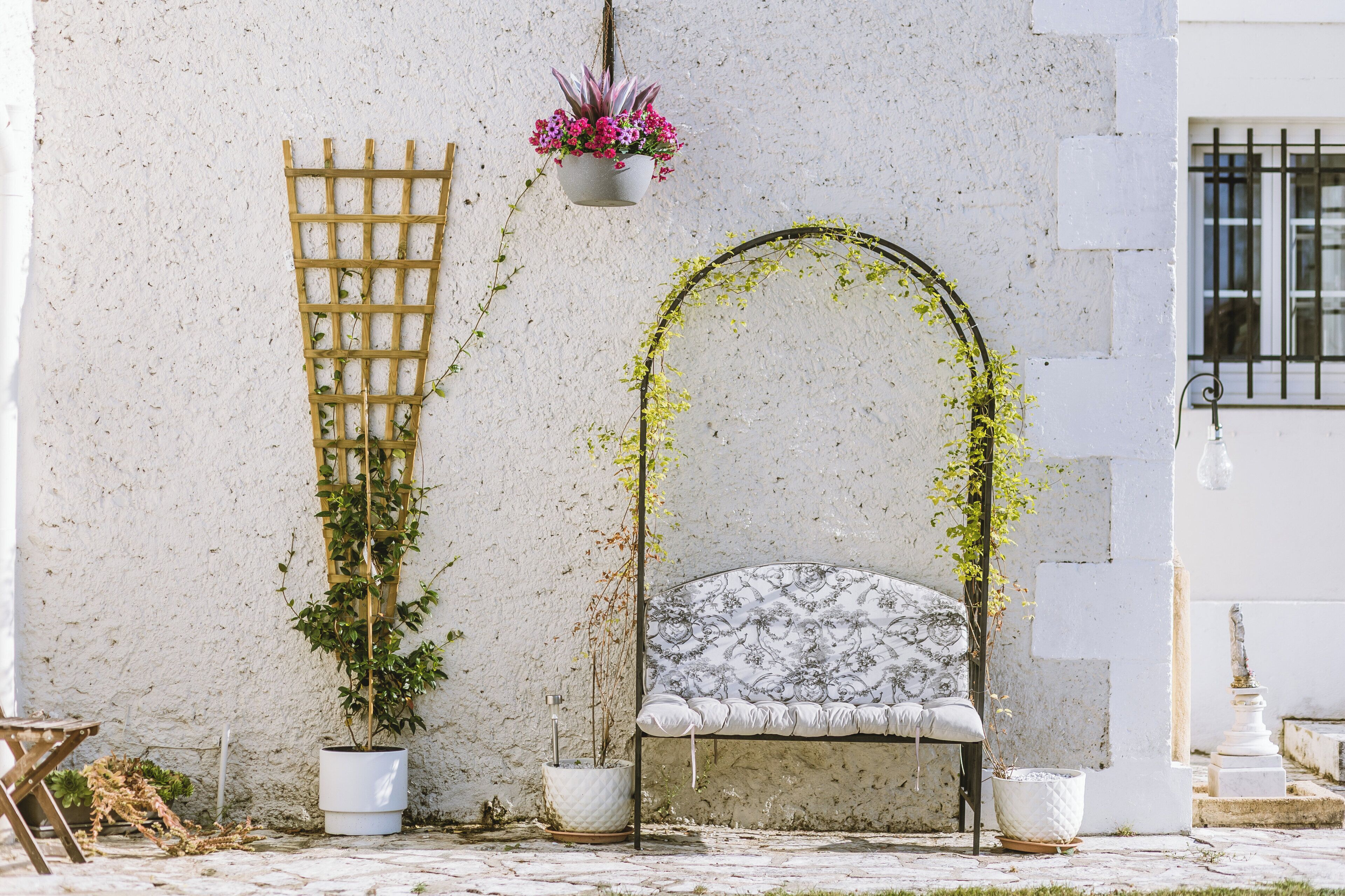 Een witte muur met houten lattenwerk, een hangende bloempot, een witte kussenbank en een plant in een pot.