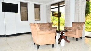 A room with two beige armchairs, a small wooden table, a flat-screen TV mounted on the wall, and a window with a view of greenery. - Hotel Twin Towers (Homa Bay)