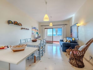 A dining area with a table, chairs, and a basket of bread. There is a sofa and wicker chairs in the living area. A television is on a stand, and a painting is on the wall. - Ocean Mood by Interhome (Porches)