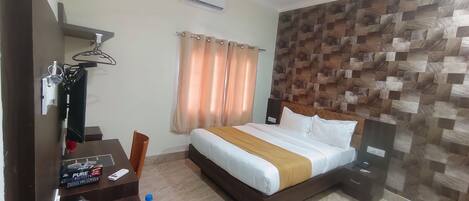 A hotel room with a bed, a desk with a computer, a chair, a wardrobe, a mirror, a ceiling fan, and a wall with a patterned design.