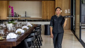 Un comedor con una larga mesa de madera dispuesta para una comida, un camarero sosteniendo una bandeja con vasos, y una cocina moderna al fondo.