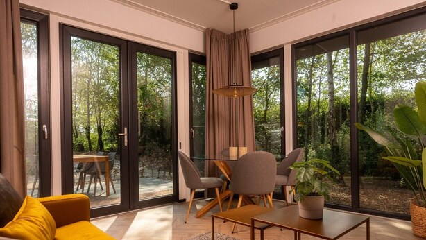 A modern living room with a sofa, a dining table with chairs, a hanging light fixture, and large glass doors leading to an outdoor area with trees. - Four-Person Bungalow in Overijssel (Zuna)