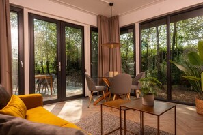 A modern living room with a sofa, a dining table with chairs, a hanging light fixture, and large glass doors leading to an outdoor area with trees. - Four-Person Bungalow in Overijssel (Zuna)