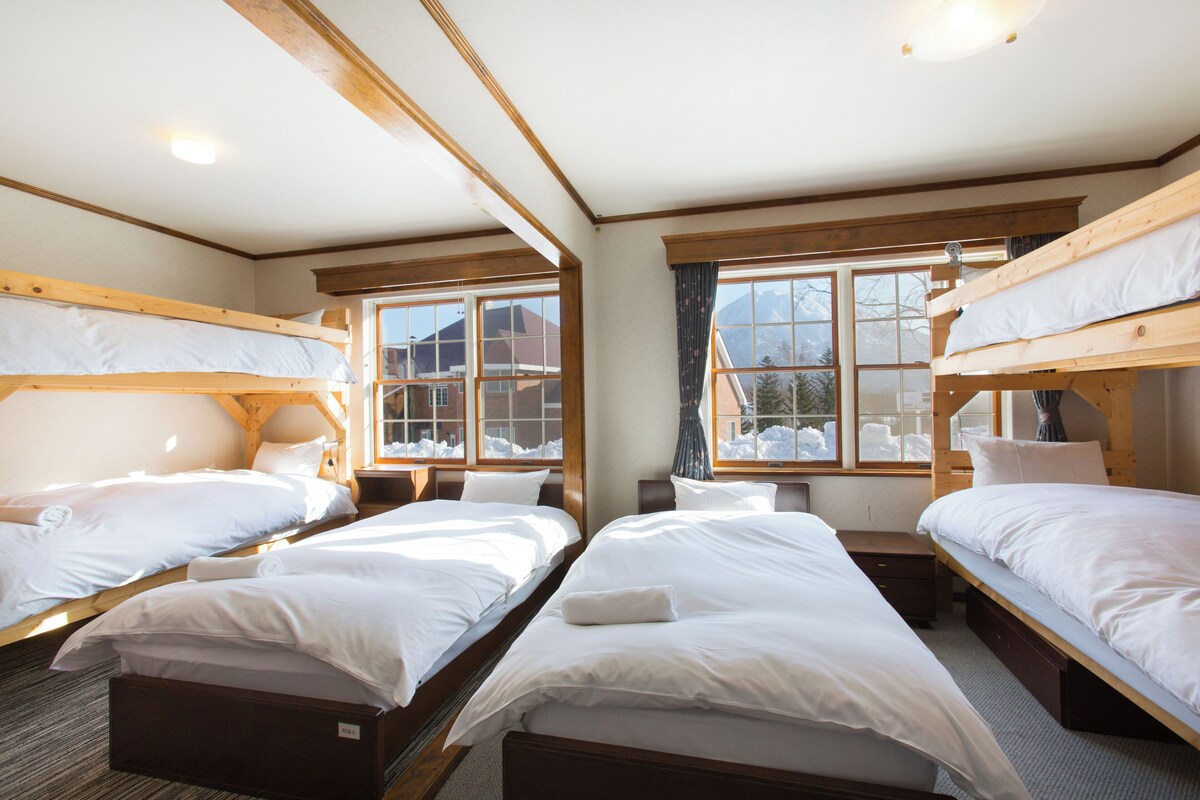 A hotel room with four bunk beds, wooden beams, large windows, and a view of snow-covered mountains.