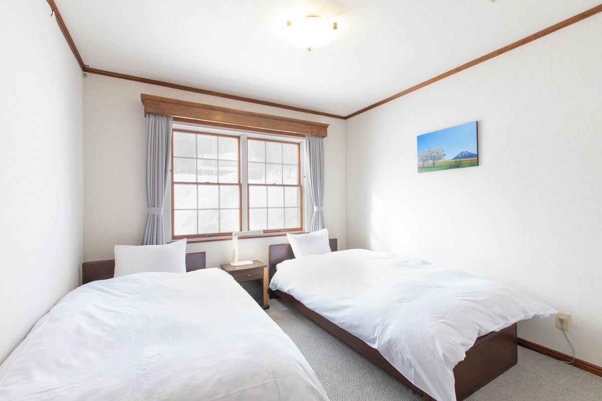 A hotel room with two single beds, a nightstand, a bedside table, a window with curtains, and a wall-mounted picture.