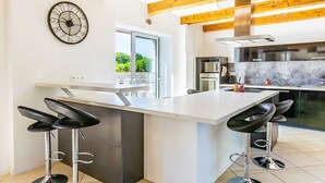 Une cuisine moderne avec un îlot central, des tabourets de bar, une grande horloge et une vue sur la verdure à l’extérieur.