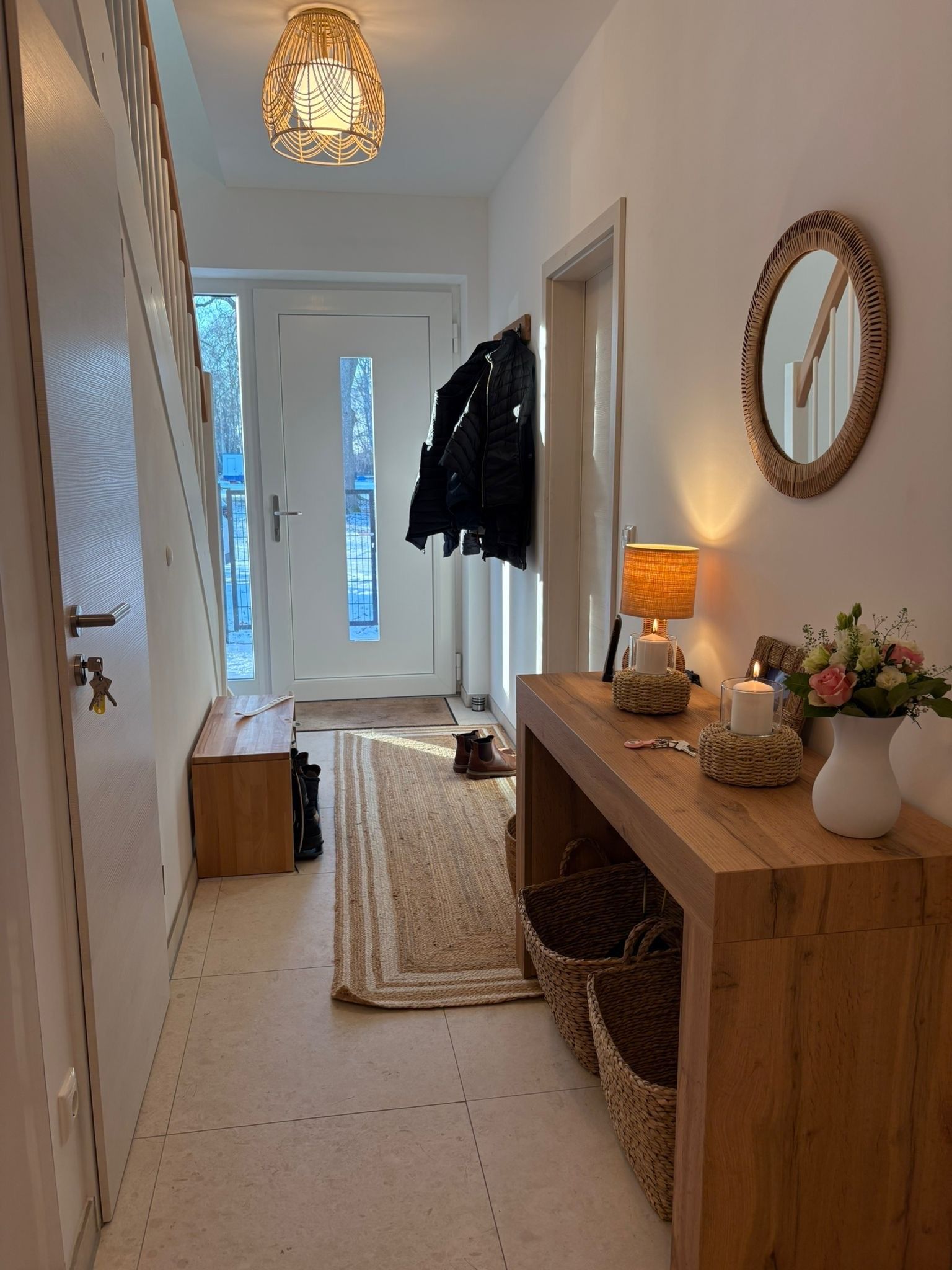 Een gang met een houten bankje, een geweven vloerkleed, een spiegel, een lamp en een vaas met bloemen.