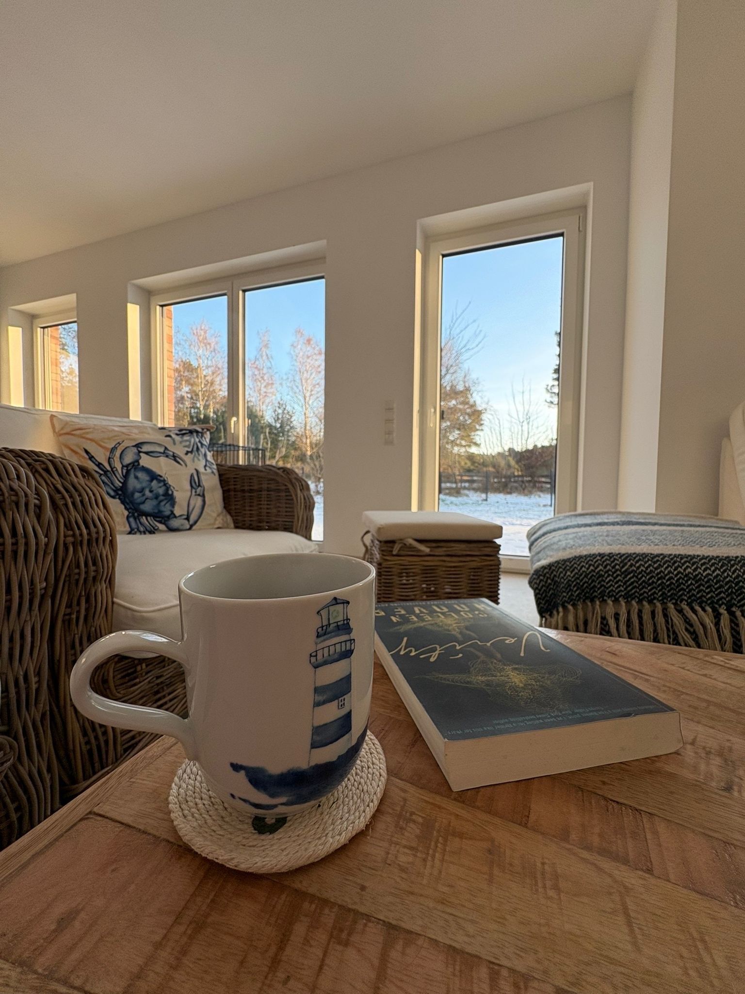 A cozy room with a large window, a wicker sofa, a white armchair, a wooden table with a mug and a book.