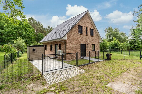 Ein einstöckiges Ziegelhaus mit schrägem Dach, umzäuntem Garten und kleiner Veranda.