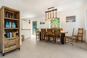 Un comedor con una mesa y sillas de madera, una estantería con libros y adornos, y vistas al exterior con vegetación.