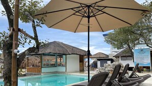 Un espace aménagé au bord de la piscine, avec des chaises longues, une structure à toit de chaume et un panneau d’affichage.