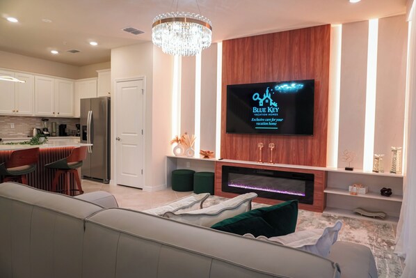 A modern kitchen with a built-in island, a dining area with a white sofa, a TV mounted on a wooden wall, and a fireplace with a stone surround. - 5BR Themed Villa · Pool · Windsor Cay · Disney (Clermont)