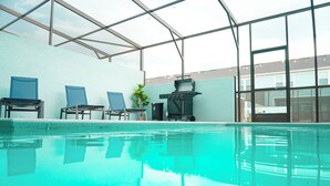 Une piscine intérieure à l’eau claire, avec des chaises bleues, un barbecue et une vue sur les bâtiments à travers de grandes fenêtres.