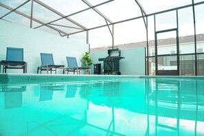 An indoor swimming pool with clear water, blue chairs, a grill, and a view of buildings through large windows. - 5BR Themed Villa · Pool · Windsor Cay · Disney (Clermont)