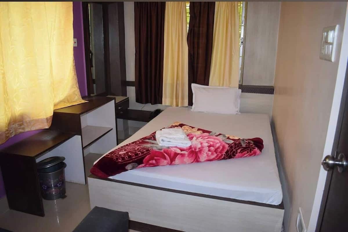 A single bed with a white mattress, a red floral blanket, and a white pillow. There is a small bedside table with a lamp, a trash bin, and a window with curtains.